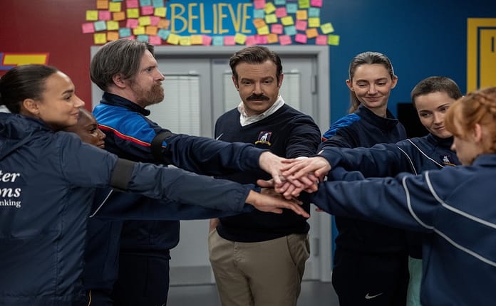 Ted Lasso, Beard, Alice CHilton and three players putting hands int he center under the believe sign with post it notes around it.